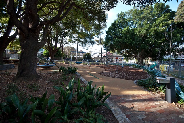 A playground with a swing set, climbing equipment, and fenced-in area surrounded by trees and a paved path.