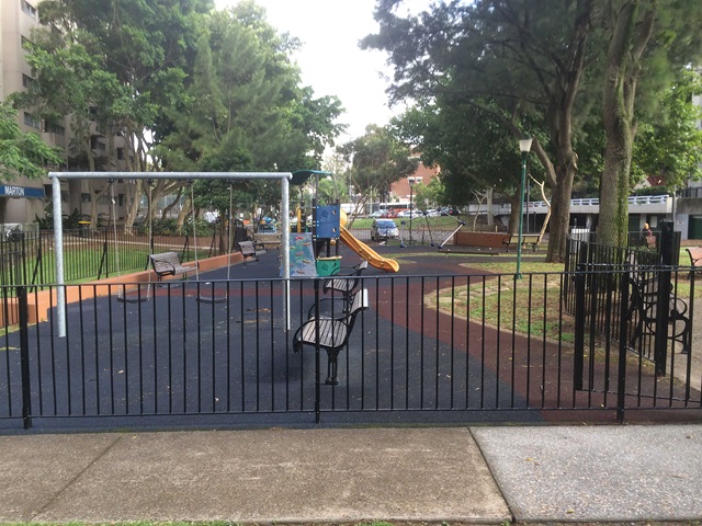 A playground with swings, a slide, benches, and trees, enclosed by a black fence.