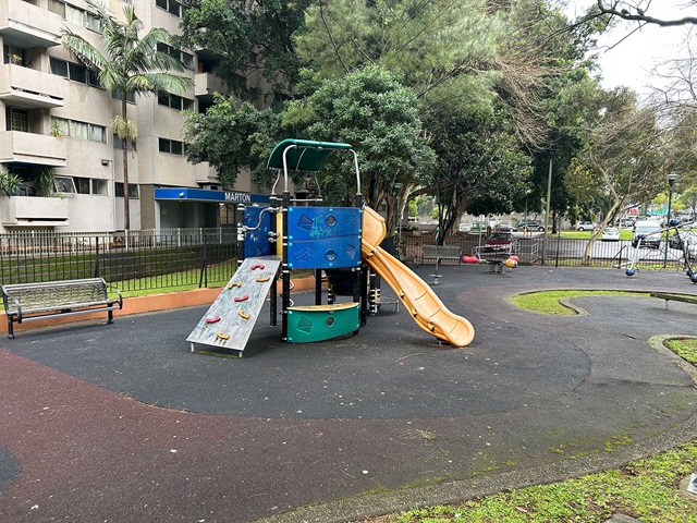 A small playground with a climbing structure, slide, and rock wall, surrounded by trees and an apartment building in the background.