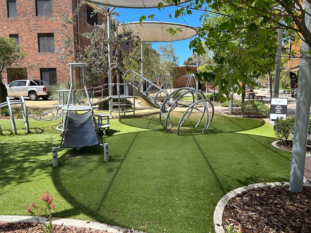 A small urban playground with swings, shaded areas, and landscaping, surrounded by tall buildings.