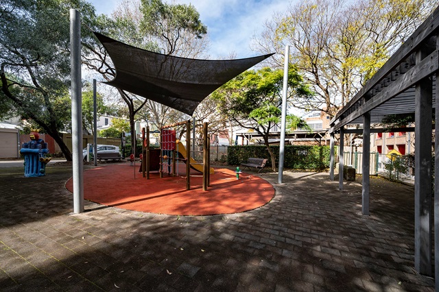 A small playground with a slide and climbing structure under a shade cover, surrounded by trees and a fence.