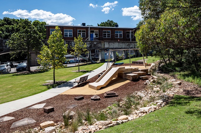 A playground with a slide and swings is surrounded by trees and grass. A brick building and parked cars are in the background under a blue sky.