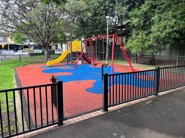A fenced playground with a slide and swings on a colorful rubber surface, surrounded by trees.