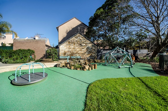 A playground with a merry-go-round, climbing structure, and stepping logs on a green rubber surface, surrounded by trees and buildings.