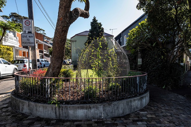 A small circular garden with metal fencing, vegetation, and a curved wire structure is set in an urban area. A sign reads "Give way to pedestrians.
