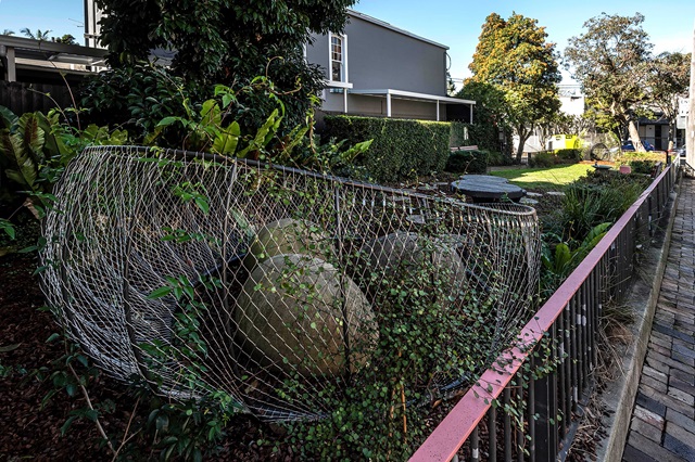 A garden with large rocks enclosed in a wire mesh, surrounded by greenery and a red railing on a sunny day.