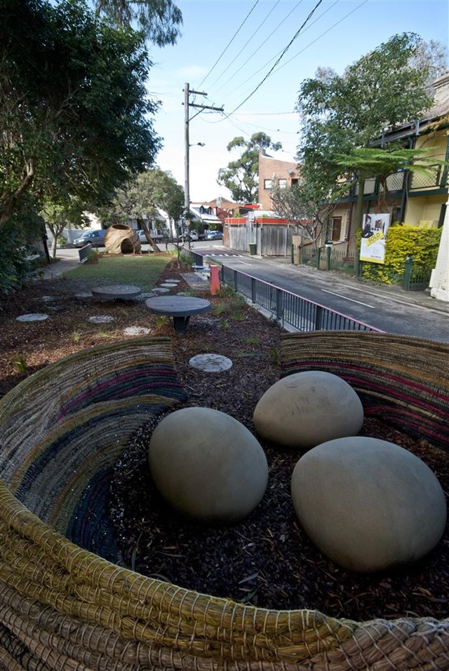 Street scene with oversized eggs in a basket sculpture on a landscaped area beside a sidewalk. Trees, a fence, and residential buildings line the street in the background.