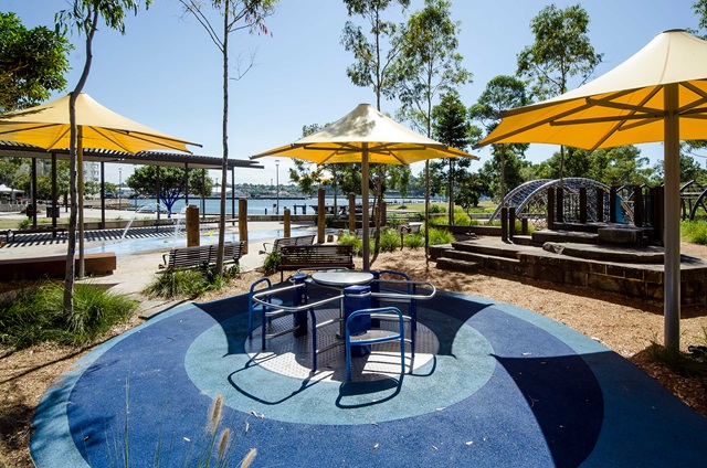 Children's playground with blue flooring, round seating, yellow umbrellas, and play equipment. Surrounded by trees, with a waterfront view in the background.