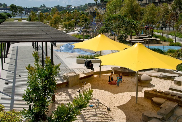 A playground with yellow shade sails covers a sandy area. A child and adult play in the sand. Trees, pathways, and seating are in the background.