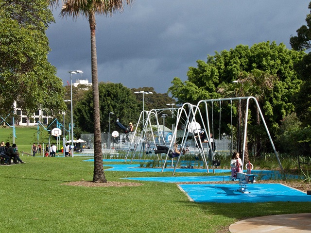 Children and adults enjoy swings and playground equipment in a park with green lawns and cloudy skies. Basketball courts are visible in the background.