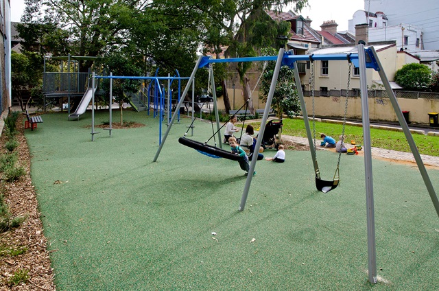 Playground with swings, a slide, and people sitting on the ground. Trees and residential buildings in the background.