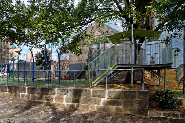 Playground with a slide, climbing structures, and safety fencing, set near a brick building and shaded by trees.
