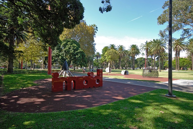 A park scene featuring a landscaped garden, palm trees, and a sculpture with the word "mod" in a shaded area.