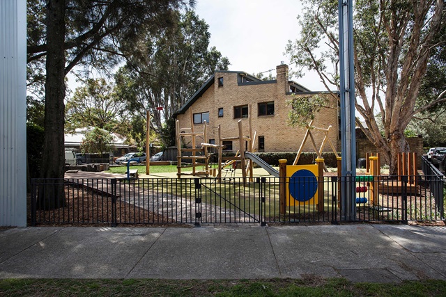 A fenced playground with slides and climbing equipment is situated in front of a large brick building, surrounded by trees.