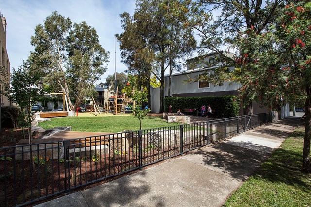 A small, fenced park with grass, trees, benches, and a play structure. A sidewalk runs alongside, and several people walk nearby.