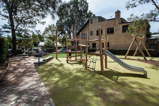 Children's playground with a wooden climbing structure, slide, and soft flooring. Surrounded by trees and near a brick building.