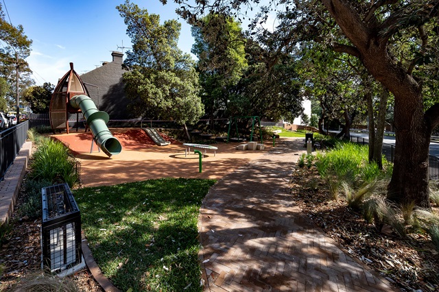 A playground features a green slide, benches, and trees, surrounded by a pathway and greenery.