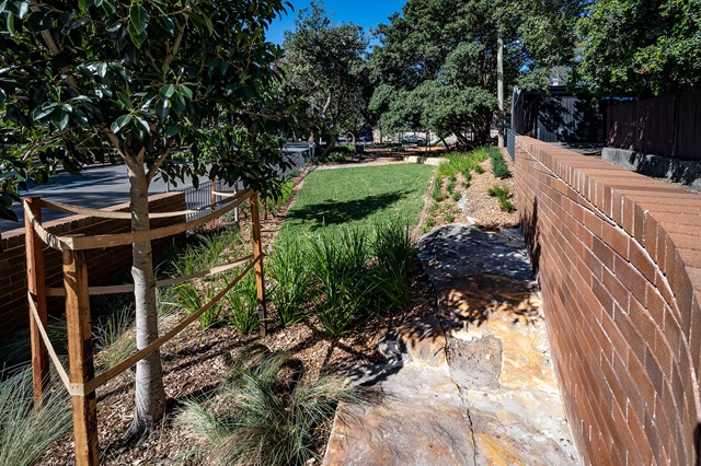 A small grass lawn bordered by a brick wall and trees, with a wooden structure supporting a young tree on the left.