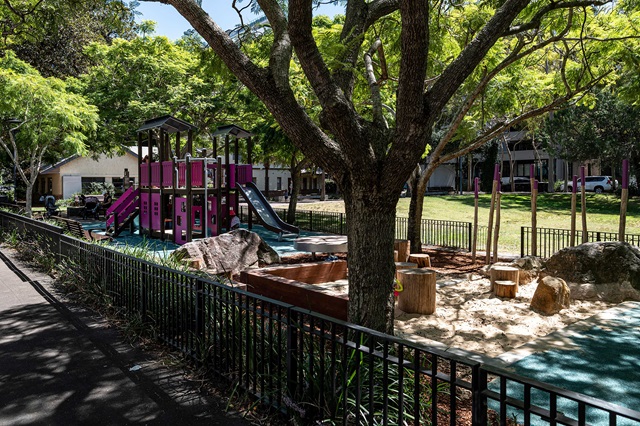 Playground with climbing structures and slides under large trees, surrounded by a black metal fence. Sand play area with rocks and a small building nearby. Pathway runs alongside the playground.