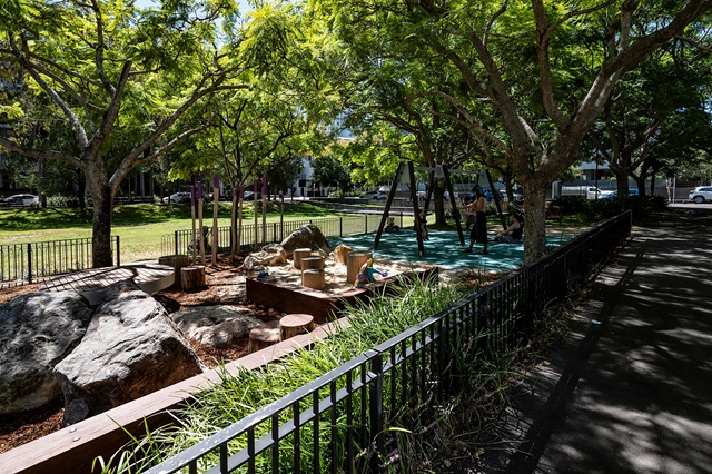 A playground surrounded by large trees, featuring swings, stepping logs, and rocks, with a pathway and fenced enclosure.