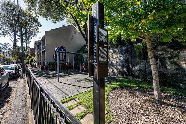 A small urban playground with a swing set and a slide, located next to residential buildings and trees, with a sign reading "Avenue Playground.