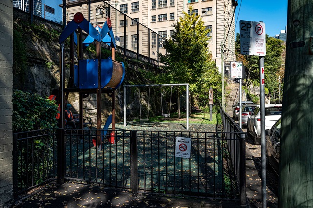 Small urban playground with a blue climbing structure and swing set, surrounded by a fence. Street signs and parked cars are visible on the nearby road.