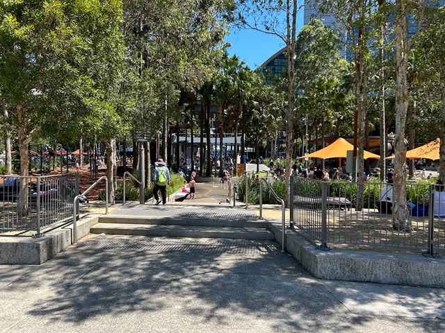 People relaxing at a park with strollers and shaded seating areas. Trees and playground equipment are visible under a clear blue sky.