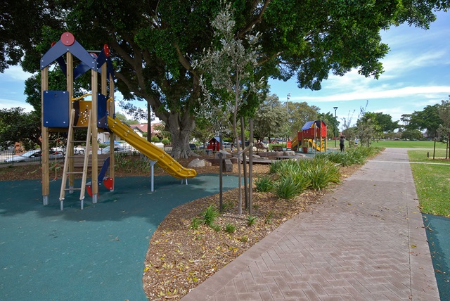 Playground with a slide, climbing structures, and trees, next to a paved path on a sunny day.