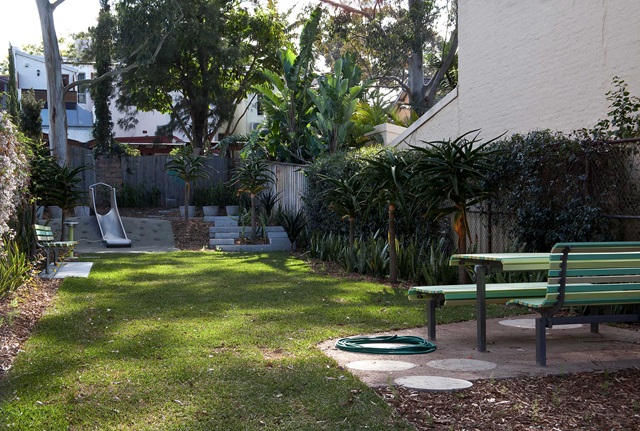 A backyard with green grass, two benches, a hose on the ground, and a slide at the rear surrounded by trees and plants.