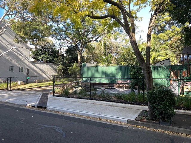 A fenced park area with trees and greenery sits adjacent to a street. There is a bench and pathway visible inside.