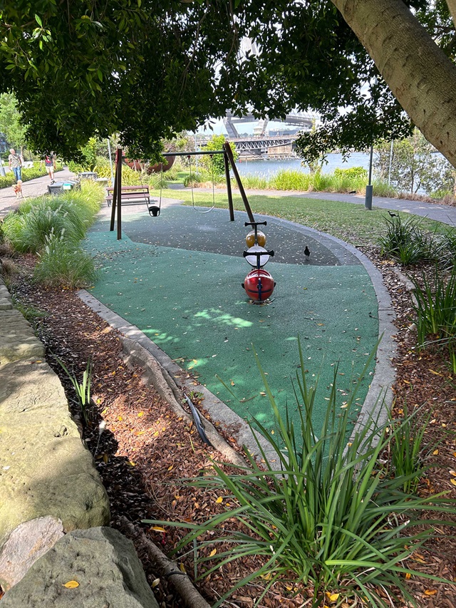 A swing set with two swings, one for toddlers, stands empty in a park surrounded by grass, trees, and a bench.