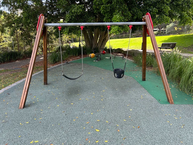 A small playground with swings and a red spring ride sits under a large tree. A path curves around the play area, and a body of water is visible in the background.