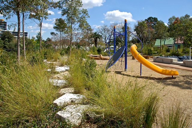A playground with a slide and climbing structure surrounded by tall grass and trees on a sunny day.