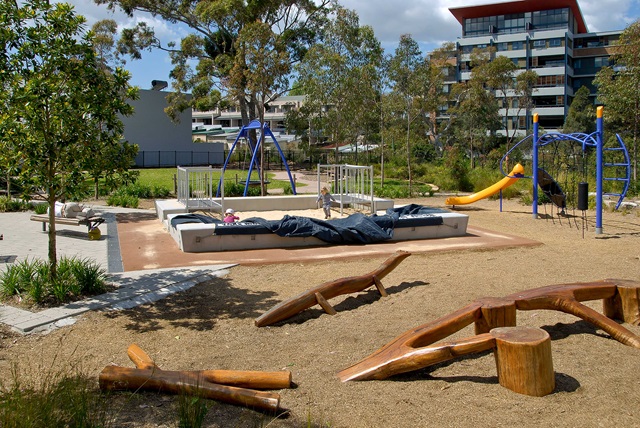 A playground with swings, a slide, climbing structures, and logs for seating. Trees and apartment buildings are visible in the background.