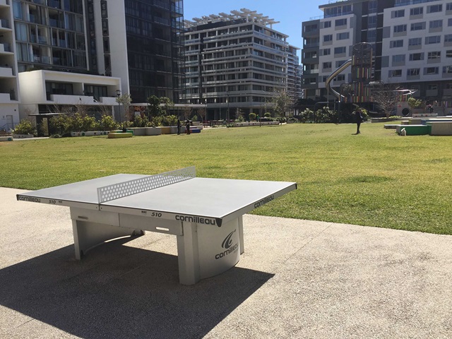 Concrete ping pong table on a sunny outdoor plaza, surrounded by modern apartment buildings and a grassy lawn.