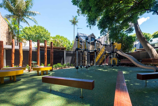 Playground with climbing equipment, slides, and benches on a sunny day, surrounded by trees and a wooden fence.