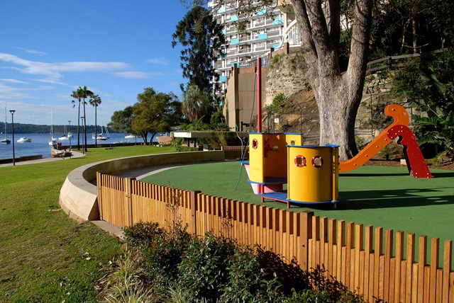semi enclosed play equipment that resembles a boat. Trees, building and a family watching a kid glide down the slide in the background.