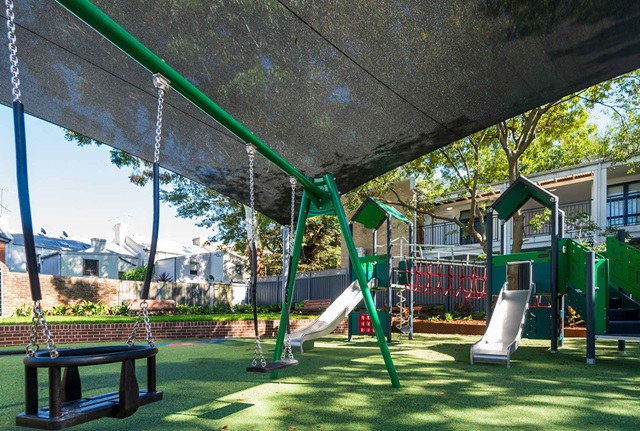A playground with swings, slides, and climbing equipment on a grassy area, shaded by a black canopy. Trees and a building are in the background.