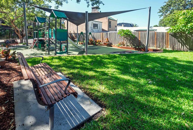 Children's playground with slides and climbing structures, a shaded area, and a wooden bench on a grassy lawn. Fence and trees in the background.
