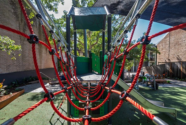Playground with red rope bridge, green slide, and climbing structure under a shade canopy. Trees and buildings surround the area.