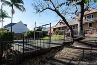 Outdoor playground area with a climbing frame and slide surrounded by a metal fence. Nearby are houses with sloped roofs and trees with bare branches.