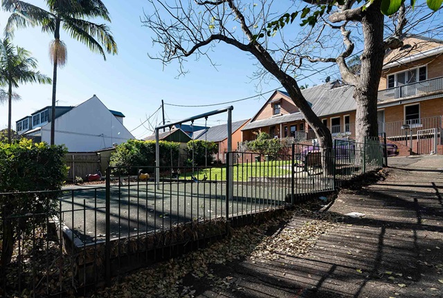 Outdoor playground area with a climbing frame and slide surrounded by a metal fence. Nearby are houses with sloped roofs and trees with bare branches.