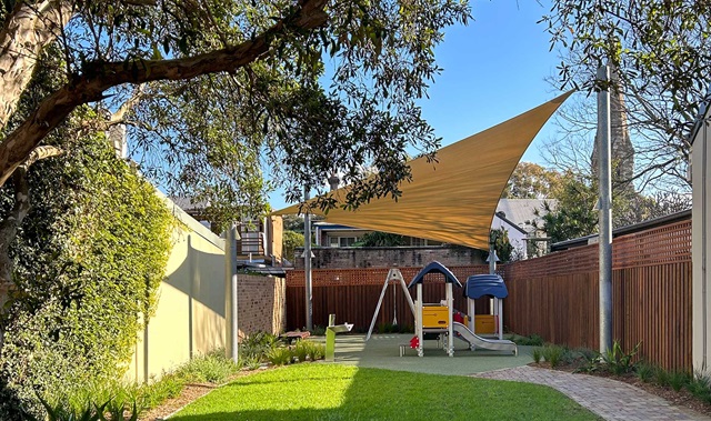 Path, lawn and plantings, in the background children’s play area with a fort, slide and shadecloth.