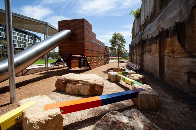 detail of steel shade structure and enclosed steel tube slide attached to a wooden enclosed playground sculpture. Sturdy stones connected with colourful beams.
