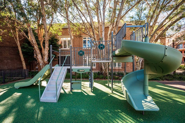Fenced play area with a curly slide, straight slide, fire pole and shopfront. Trees in the background.