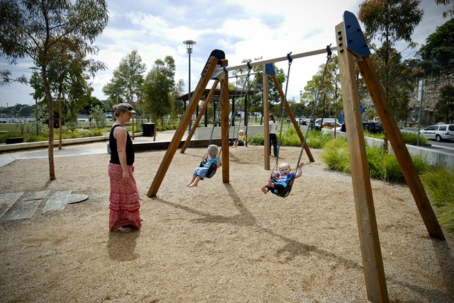 2 children playing on swings with sand as the flooring and with surrounding shrubbery in Pirrama Park Playground.