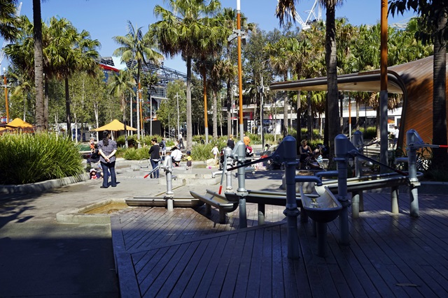 View of water play pumps in the foreground as families enjoy the playground at Tumbalong Park