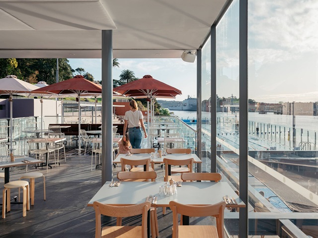 Tables and chairs in an undercover portion of the cafe on a sunny day. Shade umbrellas and outdoor area in the background. A view of the pool and Sydney Harbour on the right.