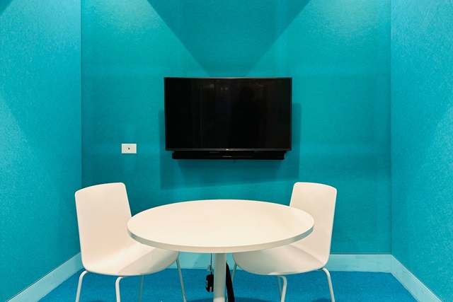 A small meeting room with light blue walls and carpet. The room contains a white table and 2 white chairs, and a screen hangs on the back wall.