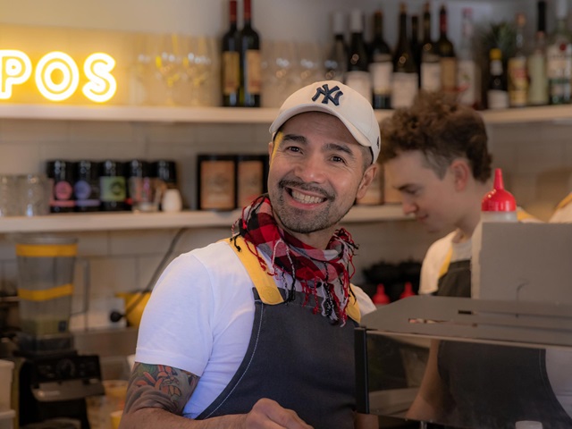 Employee wearing an apron, scarf and cap operates a coffee machine and smiles towards the camera. wine and crockery on shelves, another employee and a yellow neon sign displays the letters pos in the background.
